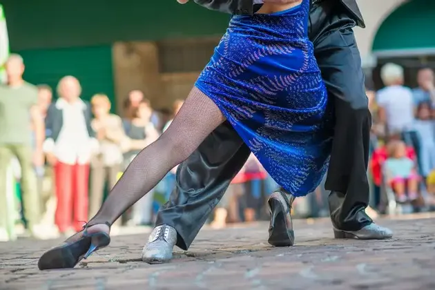 Gros plan sur les jambes d'un couple dansant le tango sur une rue pavée, avec des spectateurs flous en arrière-plan.