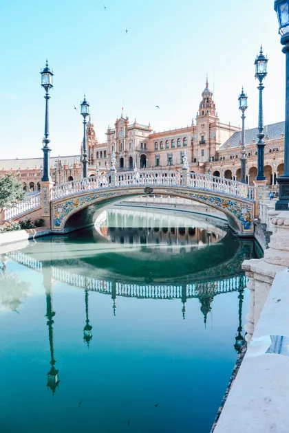 Un ponte piastrellato e riccamente decorato si inarca su un placido canale, riflettendosi nitidamente nell'acqua davanti a un maestoso edificio.