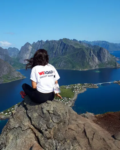 Eine Person mit einem WeRoad Group Leader T-Shirt sitzt auf einer felsigen Klippe und blickt auf einen weiten, von Bergen umgebenen Fjord.