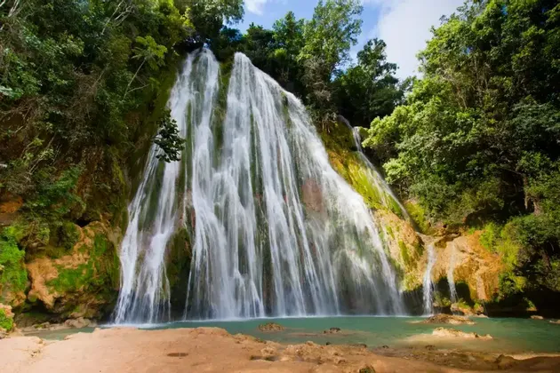 Una vasta cascata scende da una parete rocciosa ricoperta di muschio in una pozza turchese, circondata da una fitta giungla.