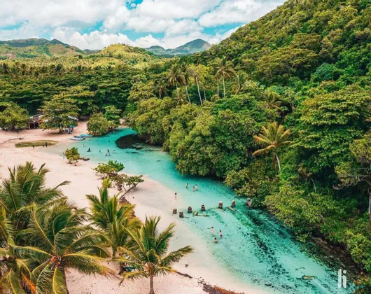 Veduta aerea di persone che nuotano nelle limpide acque turchesi di una laguna, delimitata da una spiaggia di sabbia bianca e da una fitta foresta tropicale.