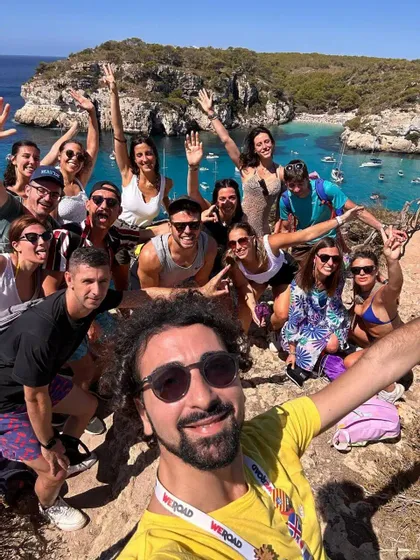Un uomo si fa un selfie di un viaggio di gruppo WeRoad su una scogliera che si affaccia su una baia turchese piena di barche.