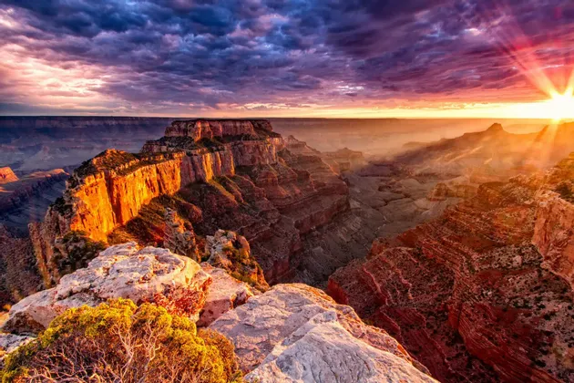 Un vasto paesaggio di canyon illuminato dal sole al tramonto, con raggi di luce che brillano attraverso nuvole viola.