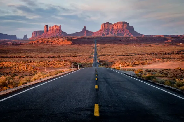 Una lunga strada dritta attraversa un paesaggio desertico arido verso imponenti formazioni rocciose rosse al tramonto.
