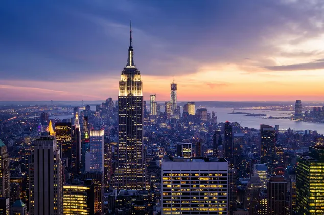 L'Empire State Building e una densa skyline cittadina sono illuminati contro un cielo colorato al tramonto sull'acqua.