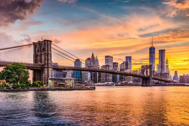 Uno skyline cittadino con grattacieli e un grande ponte sospeso su un fiume, durante un tramonto colorato con nuvole arancioni e viola.