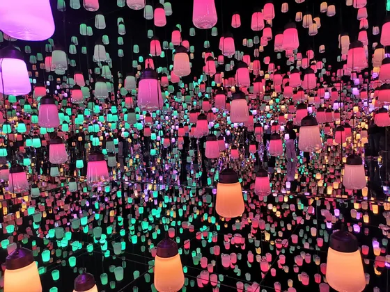 Una habitación llena de cientos de farolillos colgantes que brillan en tonos rosa y aguamarina, reflejados infinitamente por paredes con espejos.