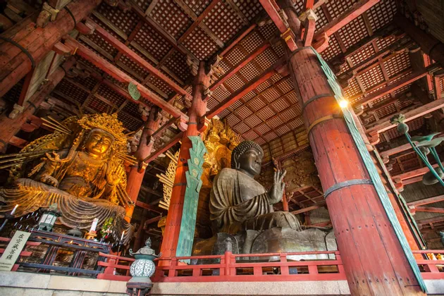 Dos grandes estatuas de Buda sentadas, una dorada y otra de bronce, dentro de un salón de madera tradicional sostenido por grandes pilares rojos.