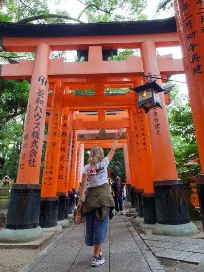 Une femme en t-shirt WeRoad Group Leader marche le bras levé dans une allée de portes traditionnelles oranges.