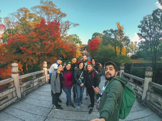 Un groupe WeRoad en voyage prend un selfie sur un pont de pierre entouré d'arbres aux couleurs d'automne éclatantes.