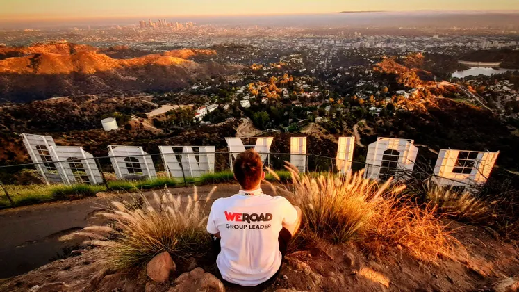 Un Group Leader WeRoad è seduto su una collina dietro la scritta Hollywood, affacciato su una vasta città al tramonto.