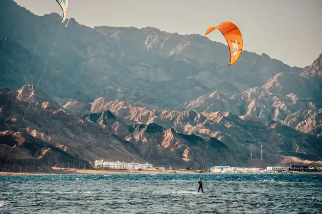 A person kitesurfs across the water with an orange kite, with a large, arid mountain range in the background.