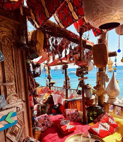 A traditionally decorated balcony with red patterned cushions and hanging lanterns overlooks a bright blue sea on a sunny day.