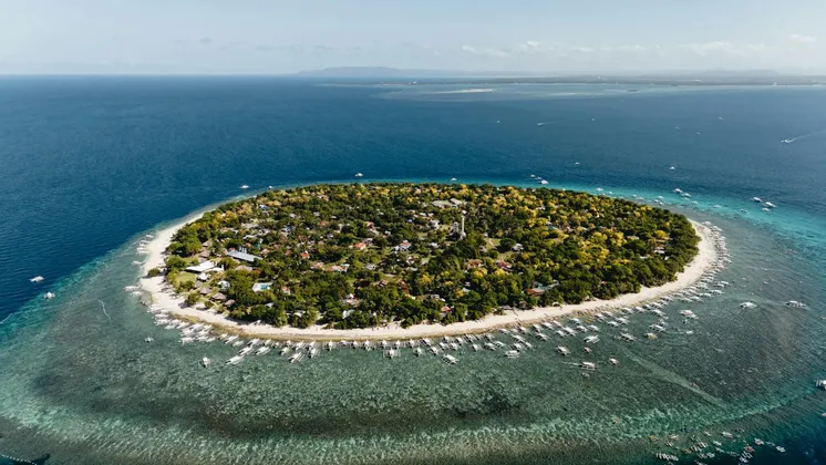Vista aerea di un'isola tropicale abitata con una spiaggia di sabbia bianca, circondata da acqua cristallina e molte barche.