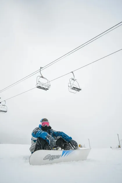 Uno snowboarder con giacca blu siede su un pendio innevato con la sua tavola sotto una seggiovia vuota.