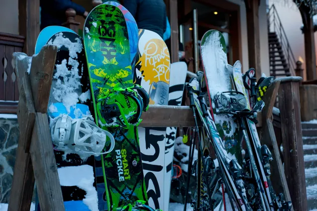 Primo piano di snowboard e sci colorati coperti di neve, posizionati su una rastrelliera di legno fuori da un rifugio.