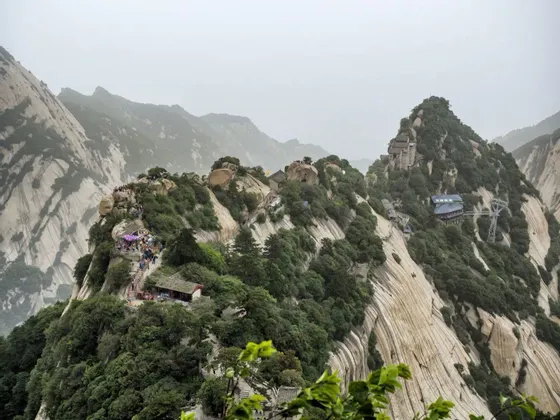 Una vista de una escarpada cordillera con picos rocosos, donde multitudes de personas caminan por un sendero entre templos y árboles verdes.