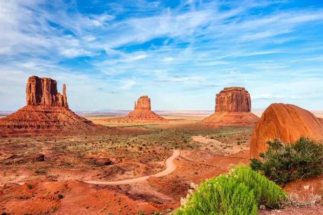Tre grandi butte di arenaria rossa si ergono in una valle desertica, con una strada sterrata tortuosa e arbusti verdi sotto un cielo blu.