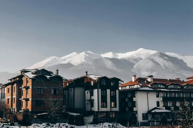 Des bâtiments de style alpin aux toits enneigés se dressent devant une vaste chaîne de montagnes enneigées sous un ciel clair.