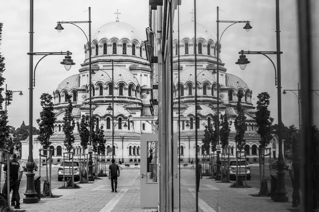 Une grande cathédrale à dôme se reflète parfaitement dans un mur de verre, créant une scène de rue symétrique en noir et blanc.
