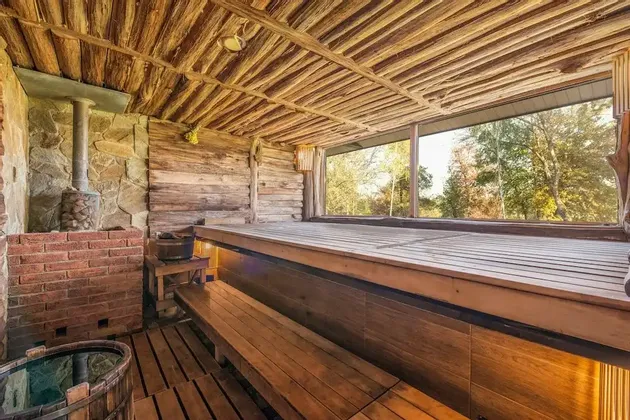 L'intérieur d'un sauna rustique en bois doté d'un poêle en pierre, de bancs étagés et d'une grande fenêtre avec vue sur la forêt.
