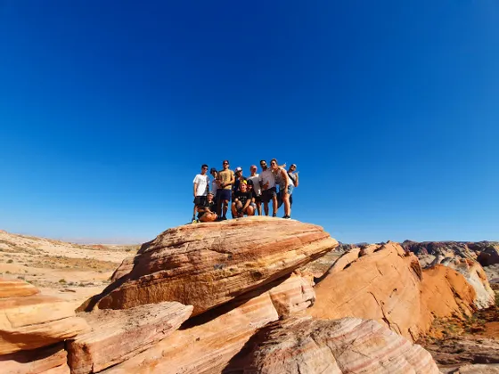 Un gruppo WeRoad in posa su una grande formazione rocciosa rossa in un paesaggio desertico sotto un cielo azzurro.