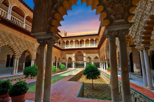 Un cortile riccamente decorato con intricati archi, colonne e una vasca riflettente centrale, osservato da sotto un arco smerlato al tramonto.