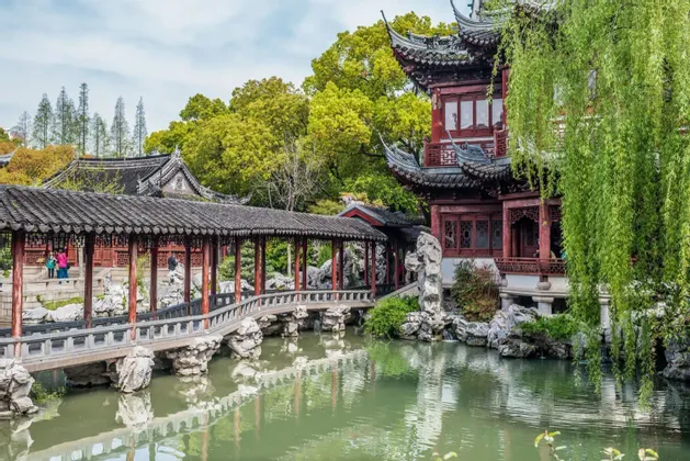 Ein traditioneller chinesischer Pavillon und eine überdachte Zickzackbrücke überqueren einen ruhigen Teich in einem üppigen klassischen Garten mit einer Trauerweide.