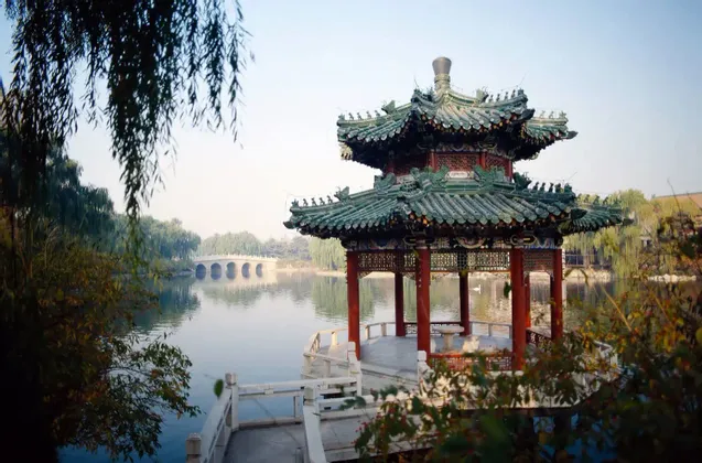 Una pagoda tradizionale con tetto di tegole verdi e pilastri rossi si erge sul bordo di un lago, con un ponte di pietra sullo sfondo.