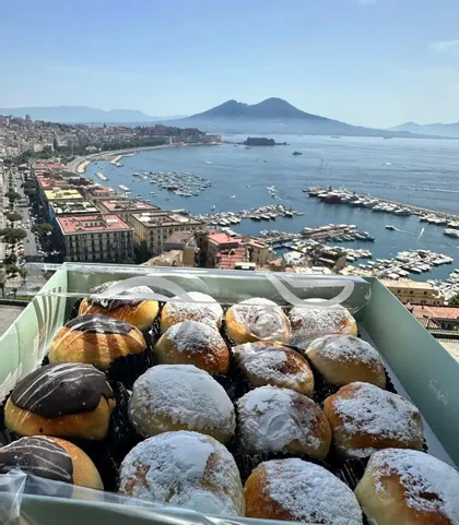 Una caja de pasteles variados con chocolate y azúcar glas se asoma a una ciudad costera con un gran puerto y una montaña a lo lejos.