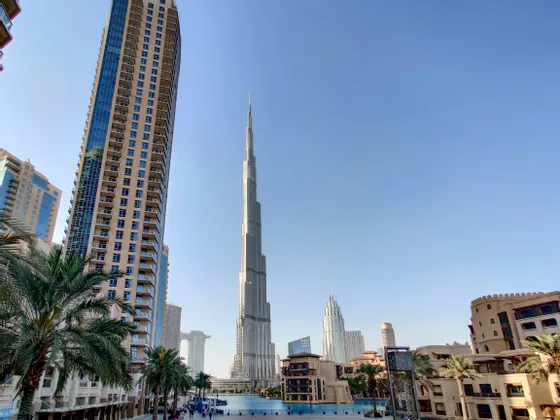 Un grattacielo alto e appuntito si erge sopra una città moderna con una grande piscina e palme in primo piano sotto un cielo sereno.