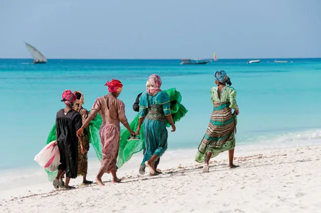 Un grupo de mujeres con coloridos trajes tradicionales camina por una playa de arena blanca junto a un océano turquesa con barcos al fondo.