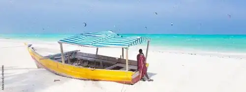 Una persona con túnica roja está junto a una barca amarilla con un toldo a rayas en una playa de arena blanca, con kitesurfistas en el océano turquesa.