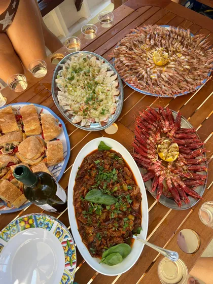Un pasto all'aperto su un tavolo di legno con vassoi di gamberetti, panini, stufato di verdure e una bottiglia di vino