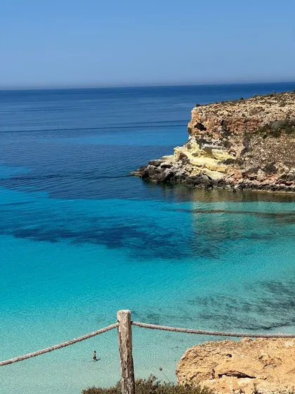 Vista dall'alto di una costa rocciosa con vivaci acque turchesi, dove una persona sta nuotando vicino a una scogliera.