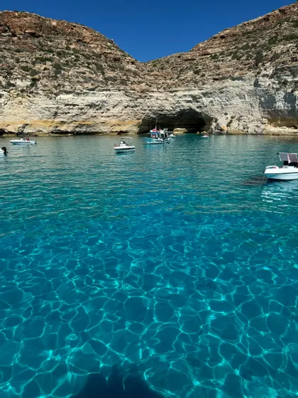 Barche su acque limpide e turchesi si avvicinano a una grotta marina alla base di una scogliera rocciosa stratificata sotto un cielo azzurro.