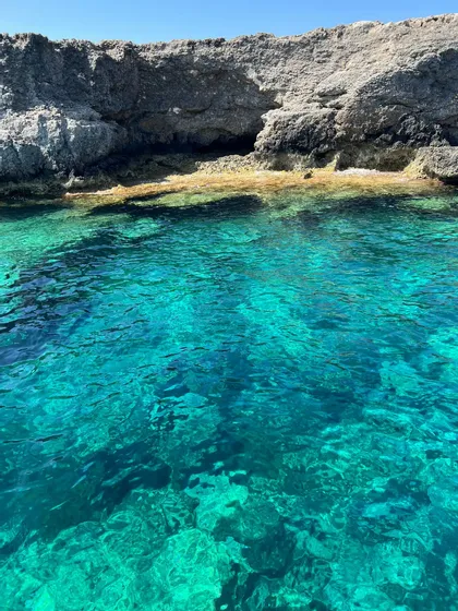 L'acqua limpida e turchese rivela il fondale roccioso ai piedi di una scogliera impervia in una giornata di sole.