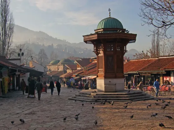 Auf einem gepflasterten Platz steht ein verzierter Holzbrunnen mit einer grünen Kuppel, während Spaziergänger an Marktständen vorbeiflanieren und Tauben sich am Boden sammeln.