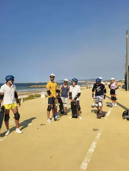 Un gruppo WeRoad in viaggio, con caschi e protezioni, si trova con gli skateboard su un sentiero asfaltato in riva al mare.