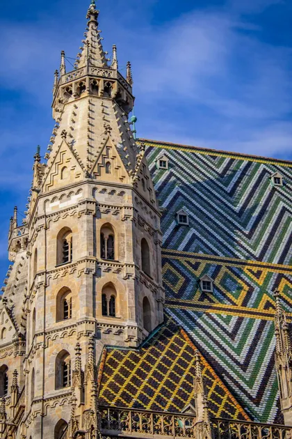 Una torre in pietra riccamente decorata di una cattedrale gotica, con un tetto di tegole colorate e a motivi, contro un cielo azzurro brillante.