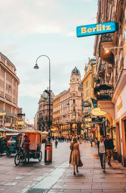 Persone camminano lungo un'ampia strada lastricata in una città, fiancheggiata da architettura ornata, negozi e un risciò parcheggiato.