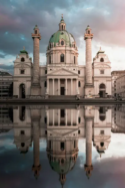 Una chiesa bianca ornata con una grande cupola verde si riflette perfettamente nell'acqua antistante sotto un cielo nuvoloso.