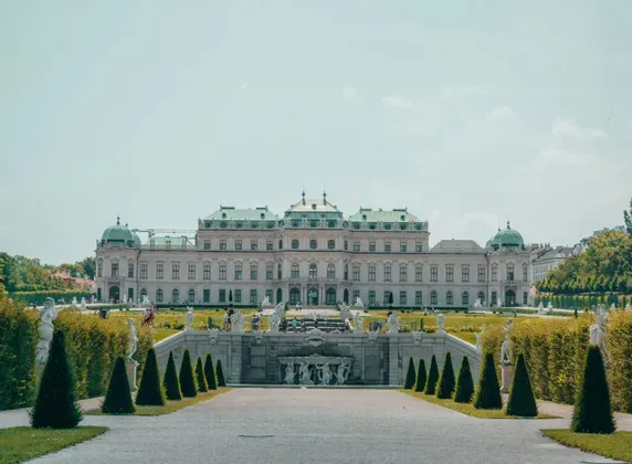 Un'ampia vista di un grande palazzo con tetto verde, situato dietro un giardino formale con una fontana e un percorso centrale.