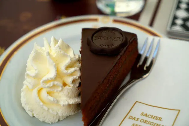 Ein Stück Sachertorte, mit Schokoladensiegel, liegt auf einem Teller neben einem Klecks Schlagsahne und einer Gabel.