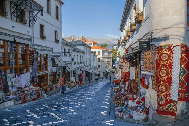 Eine enge Kopfsteinpflastergasse mit Geschäften für farbenfrohe Teppiche und Textilien und Bergen in der Ferne.