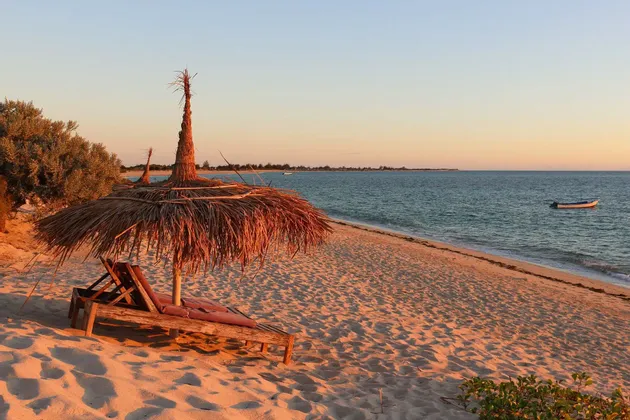 Two empty wooden lounge chairs under a thatched umbrella on a sandy beach at sunset, with a calm sea and a small boat in the background.