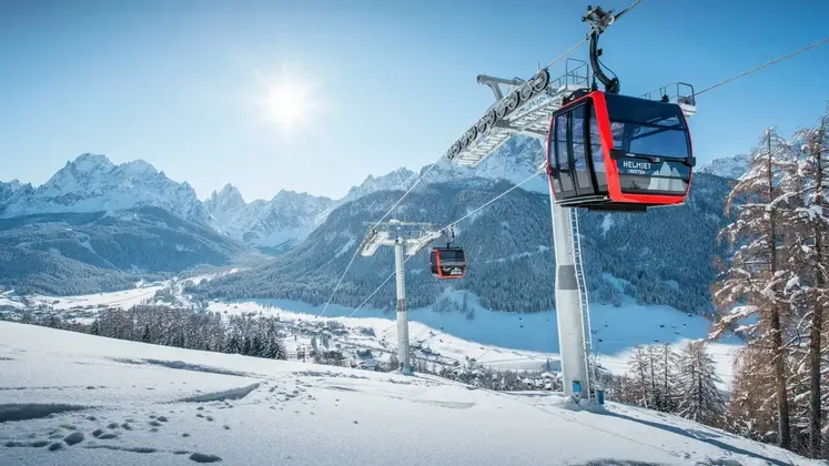 Gondole rosse salgono su una montagna innevata sotto un sole splendente, con una vasta catena montuosa coperta di neve sullo sfondo.