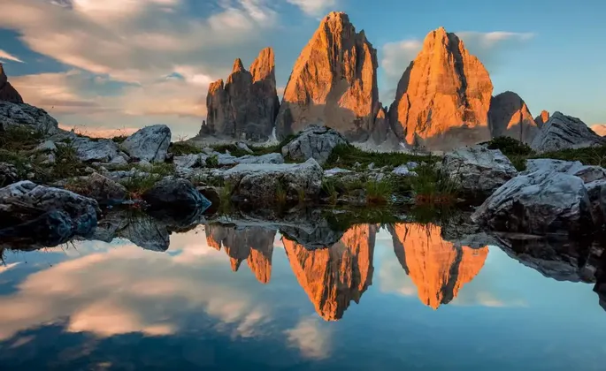 Cime montuose frastagliate illuminate dalla luce dorata si riflettono in un lago immobile con una sponda rocciosa in primo piano.