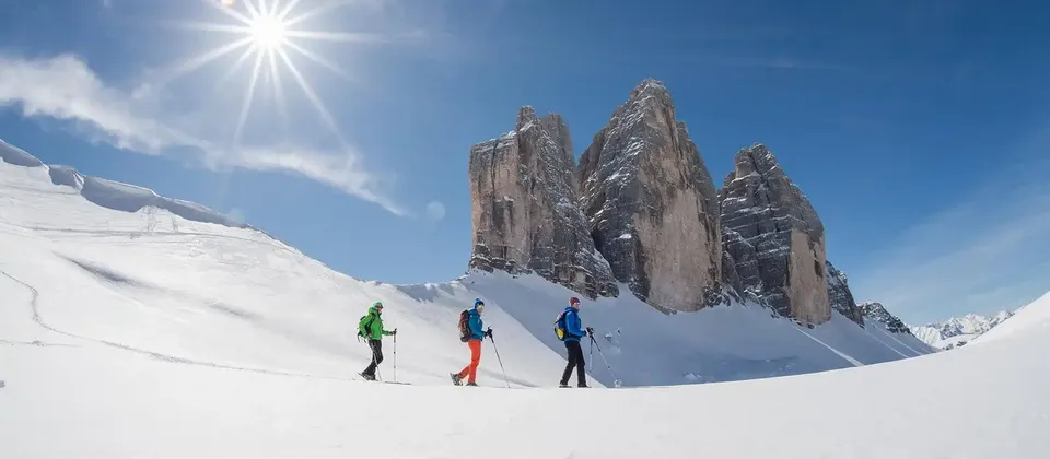 Tre persone di un viaggio di gruppo WeRoad con le ciaspole in un paesaggio montano innevato sotto un cielo limpido e soleggiato.