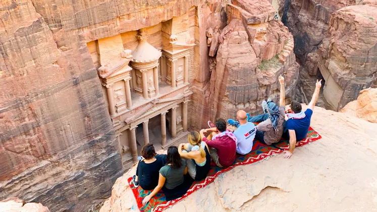 Un gruppo WeRoad si trova su un tappeto su una sporgenza di scogliera, con la vista su un grande tempio scavato in un canyon di roccia rossa.